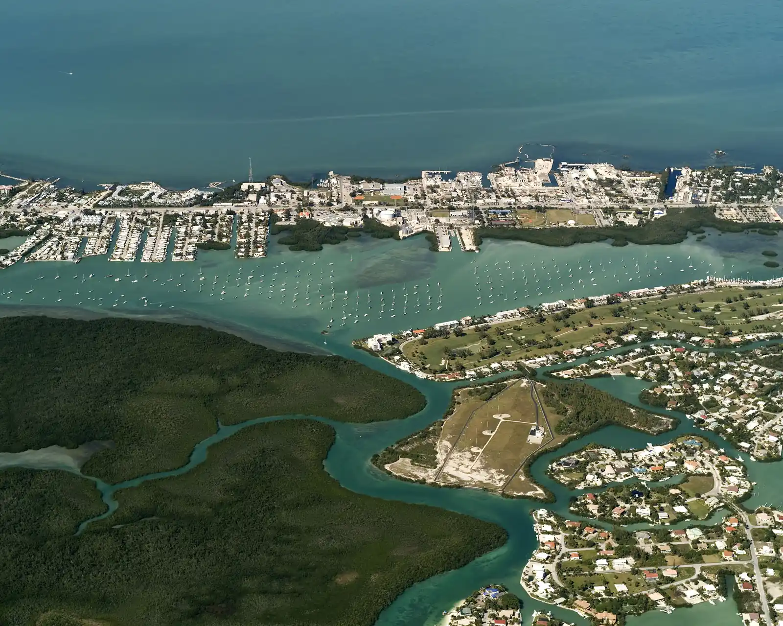 High oblique aerial looking north over Boot Key Harbor: a broad green-turquoise harbour packed with well over a hundred moored sailboats, lined on the far side by the canals, docks and buildings of Vaca Key along the Overseas Highway, with the open water of Florida Bay beyond. In the foreground is the dark-green mangrove forest of Boot Key cut by winding tidal creeks, and at right a fenced grassy field with a small building beside waterfront homes, canals and a golf course.