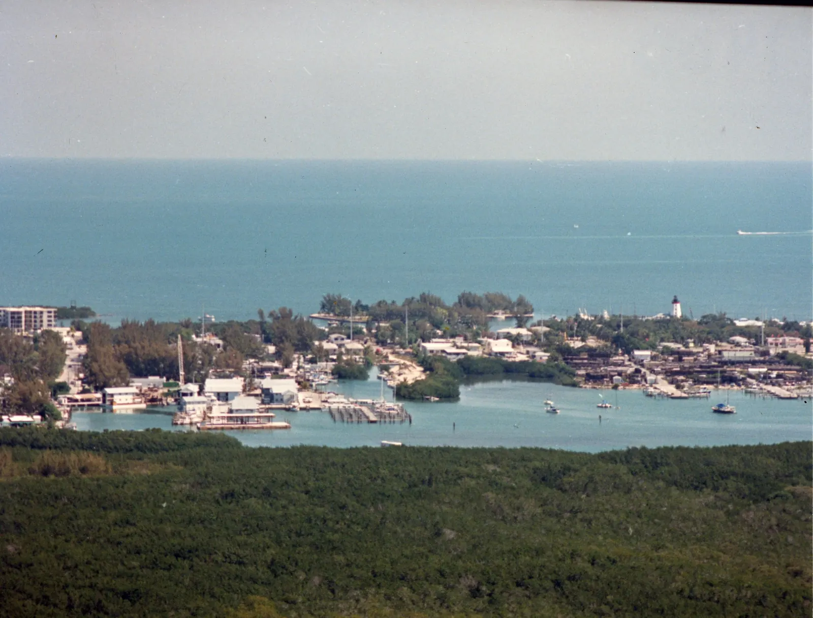 Colour aerial from 1987 across a huge expanse of dark-green mangrove in the foreground to a sheltered harbour with marinas, piers, a boatyard, docked and anchored boats and low buildings among trees. A red-and-white lighthouse-shaped tower stands near the right of the shoreline, and the open, blue-green sea stretches to a hazy horizon. Dust specks and scratches from the film are visible, with a thin dark edge at the top right.