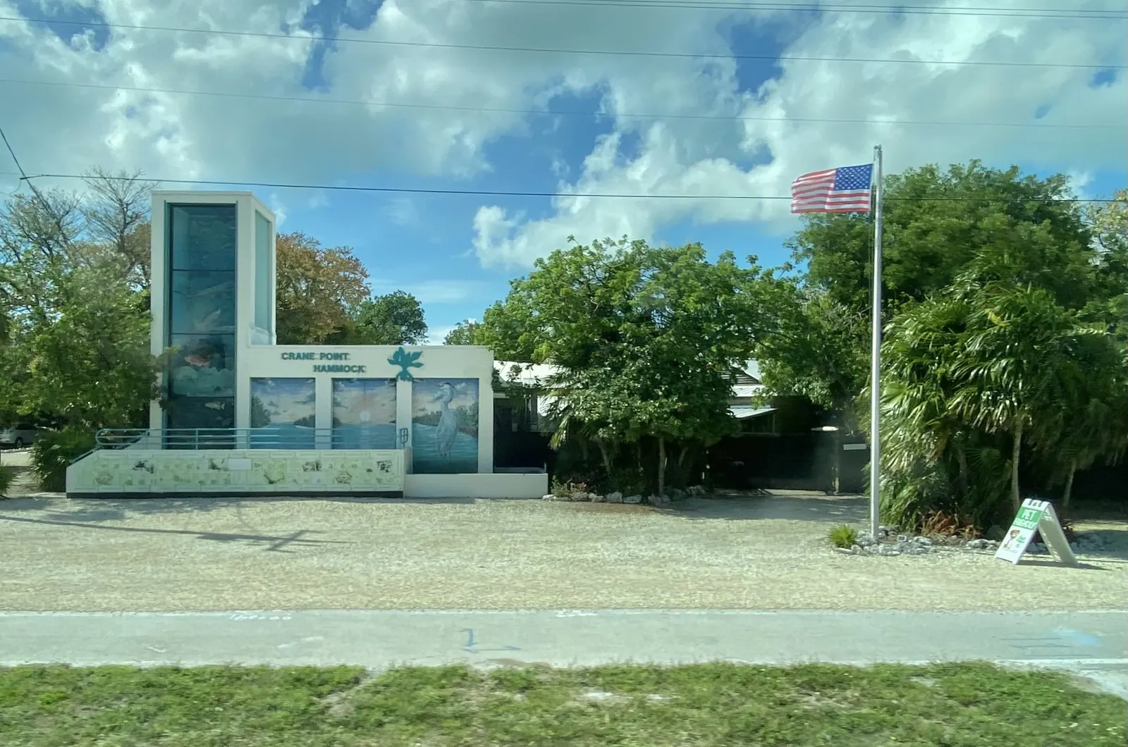 Roadside view of the Crane Point Hammock entrance building: a white structure with a tall glass tower, 'Crane Point Hammock' lettering, a teal leaf logo and murals of mangrove water, a sunset and a great blue heron. Shade trees and palms surround it, a US flag flies from a pole at right beside a small sandwich-board sign, and a paved path and grass strip run along the front under a blue sky with heavy white cloud.