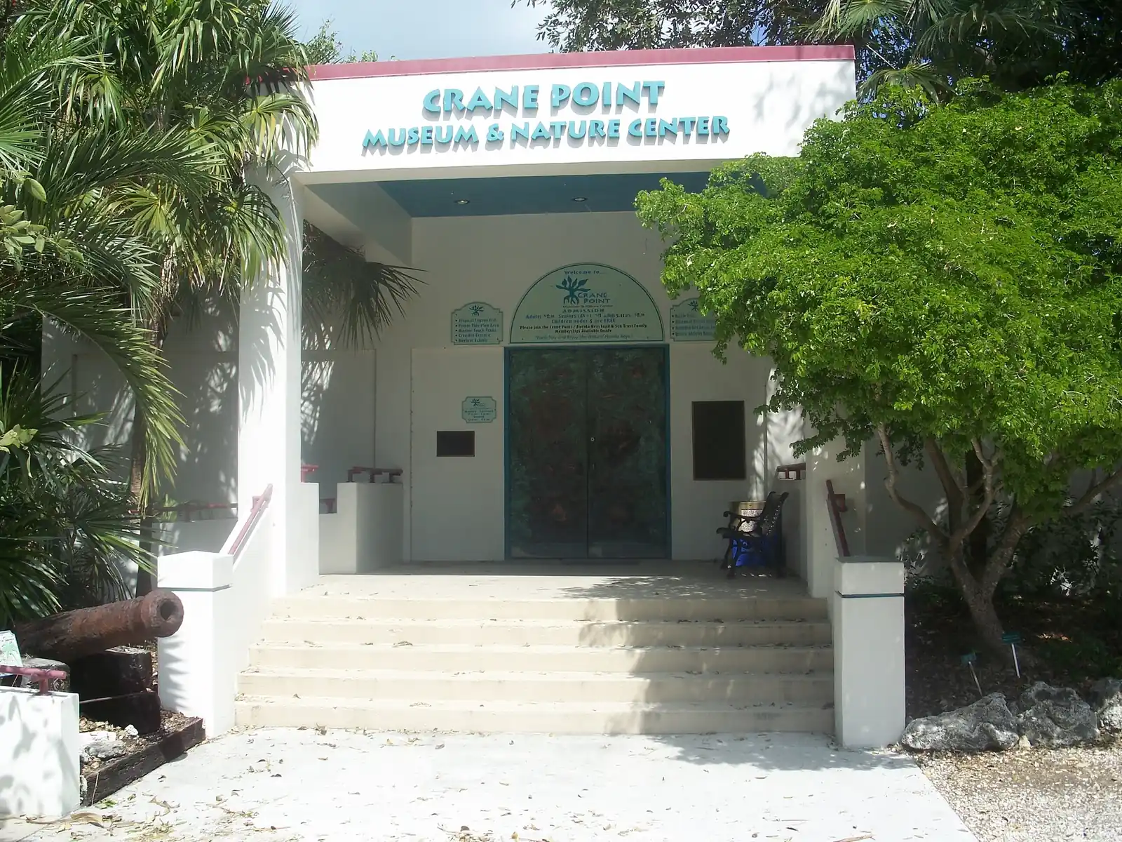 Front entrance of the Crane Point Museum & Nature Center: a white building with teal lettering under a maroon roof edge, wide concrete steps up to a covered porch, decorated double doors, an arched admission sign and small posted signs. An old rusted cannon lies at lower left; palms and a leafy tree frame the entrance, with coral-rock boulders at right.