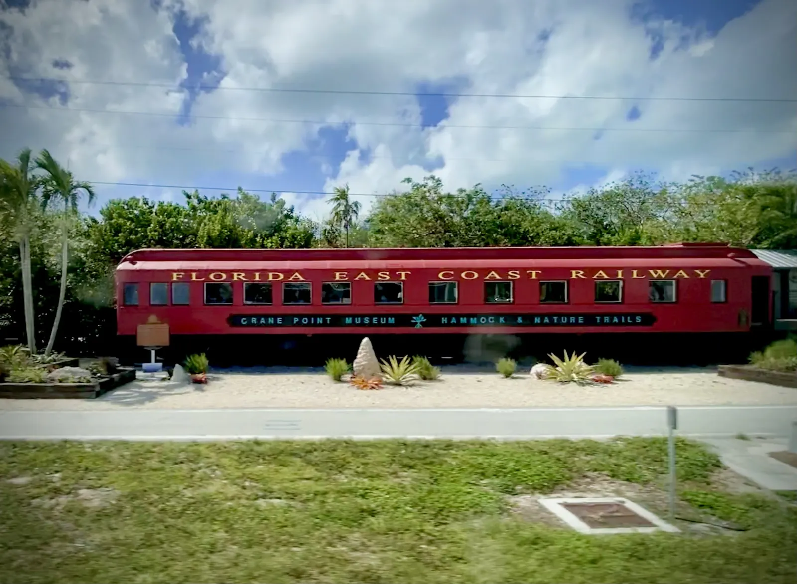 A long red railway passenger car lettered 'Florida East Coast Railway' in gold, with a banner along its side reading 'Crane Point Museum Hammock & Nature Trails', sits beside the road on a gravel bed planted with agaves and bromeliads, with a historical marker at its left end. Palms and leafy trees rise behind it under a blue sky full of white cloud; a grass verge and a path run across the foreground.