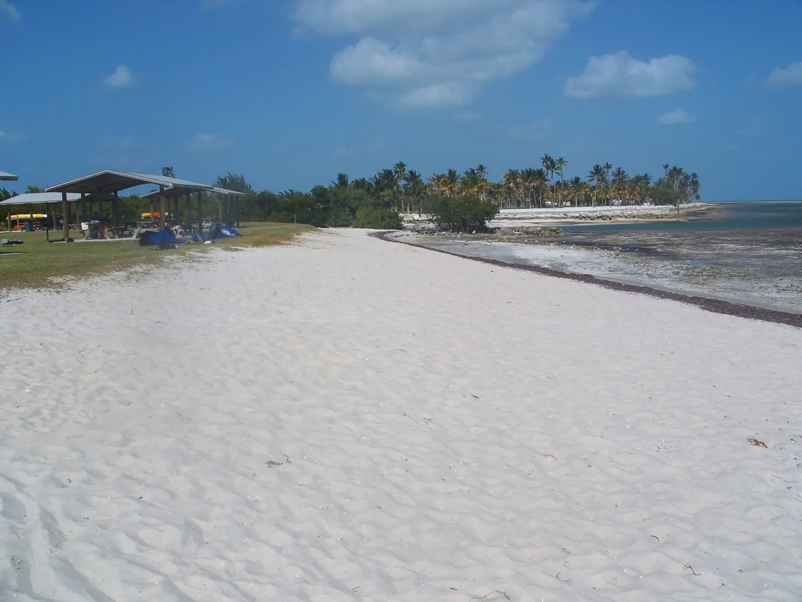A wide strip of white sand runs into the distance beside a shallow, weedy flat at low tide, with a dark line of washed-up seagrass along the edge. On the left are open-sided picnic shelters on grass, folded blue kites or gear on the sand and yellow kayaks in the background; a palm-covered point and a rock revetment sit at the far end under a blue sky with white clouds.