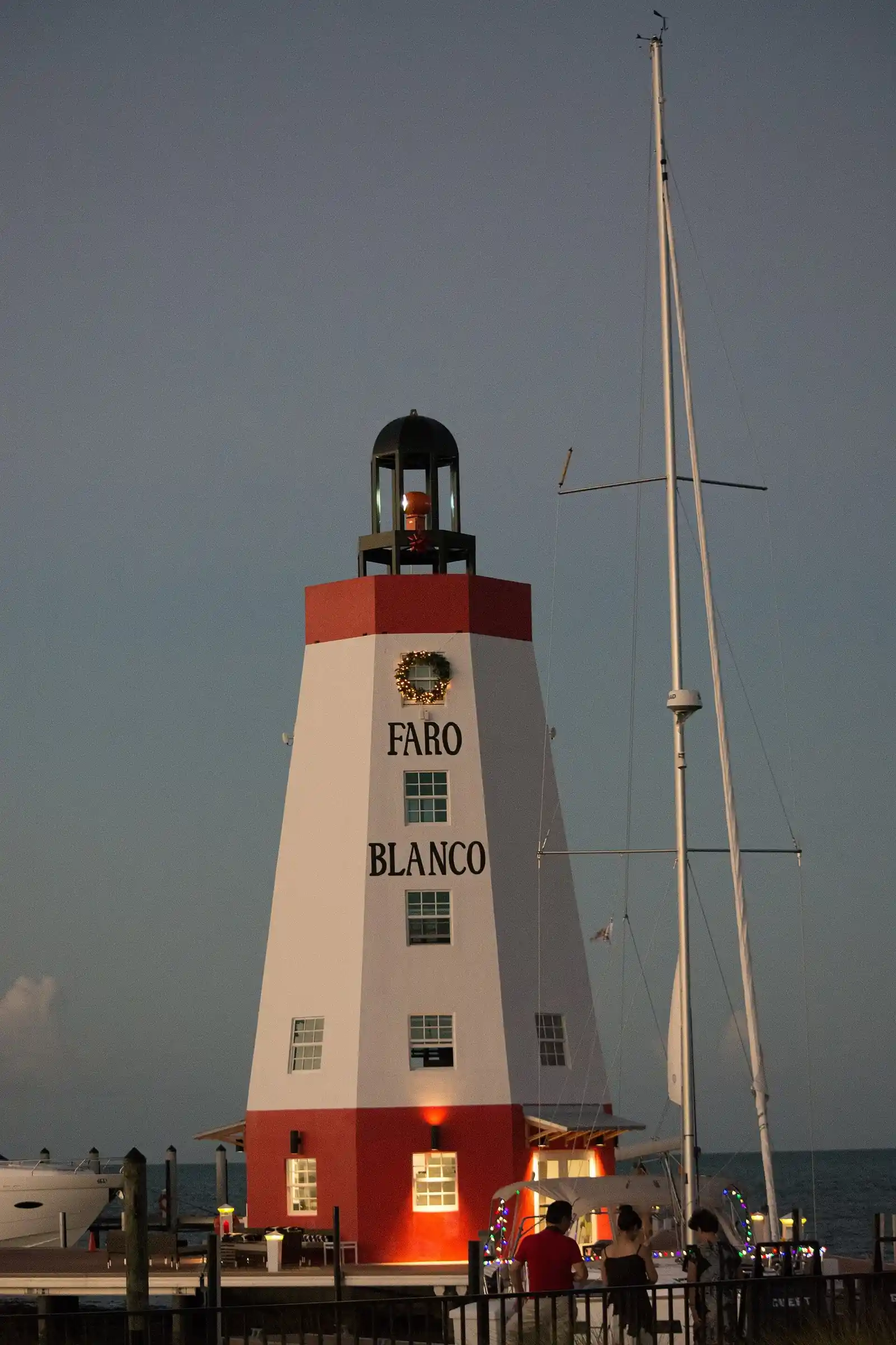 At dusk, a tapering white tower with red bands at its top and base, the words 'Faro Blanco' painted down its side and a black lantern cage on top, stands on a waterfront dock. A lit holiday wreath hangs near the top and warm lights glow at its base. To the right is a sailboat mast with rigging and a boat strung with coloured lights; four people stand on the dock in the foreground, seen from behind and in partial profile. The sky is a smooth slate grey-blue.