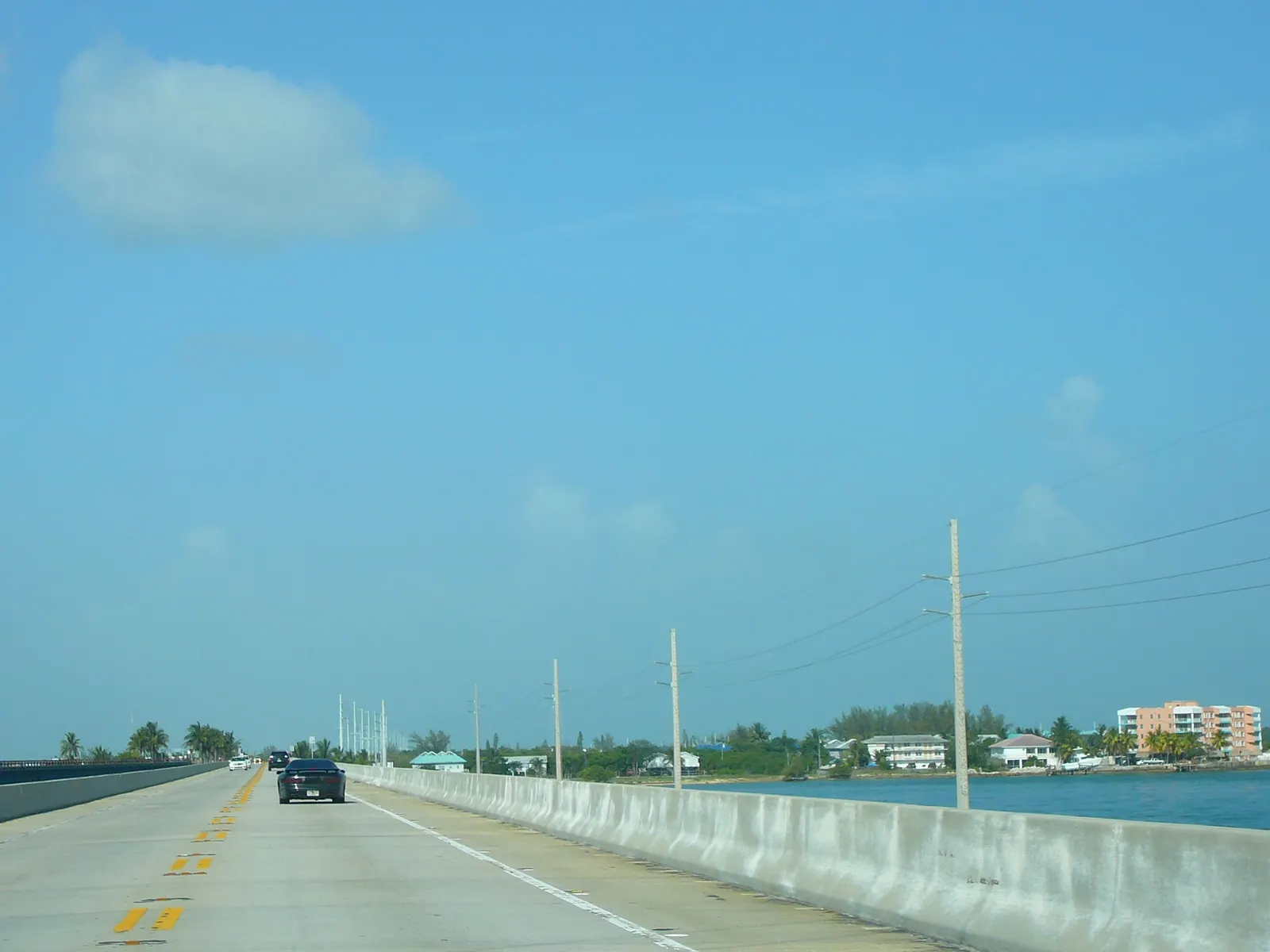 Driver's-eye view along a two-lane concrete highway bridge with a low concrete barrier at right, a few cars ahead, and a row of utility poles with wires running beside it over blue water. At the end of the bridge is a low shoreline of palms and trees with houses and a pink multi-storey apartment building at right, and at far left the deck of a parallel older bridge. The upper two-thirds is hazy blue sky with one soft cloud.