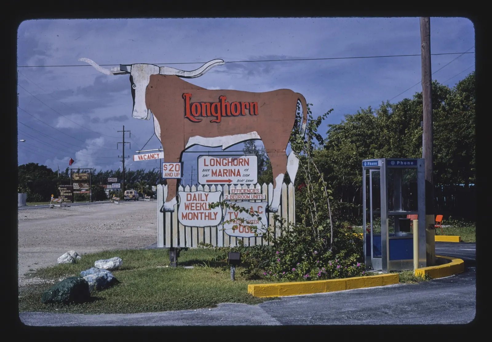 1985 colour slide of a big cut-out roadside sign shaped like a longhorn steer, lettered 'Longhorn', with smaller signs below on a picket fence: 'Longhorn Marina - beer, bait, ice, soda, boat ramp', 'Vacancy', '$20 and up', 'Daily Weekly Monthly', 'Efficiencies, 2 bedroom units' and 'TV, A/C, fresh water pool'. A phone booth stands at right beside yellow kerbing and flowering shrubs; behind are a gravel road with more business signs (including 'The Diving Site' and 'Coral Lagoon Resort'), utility poles, trees and a stormy blue sky. The slide's black border frames the image.