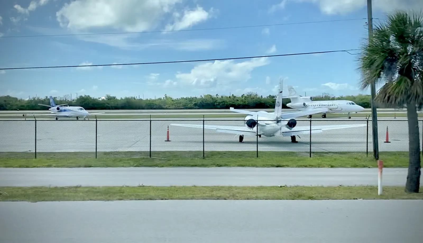Looking across a road and a grassy verge, through a chain-link fence, at the apron of Florida Keys Marathon Airport: a white twin-engine business jet seen from behind at centre, flanked by orange traffic cones, with two more white business jets parked beyond it at left and right. A low line of green trees sits behind the airfield under a blue sky with white cloud; a palm and a utility pole stand at right.