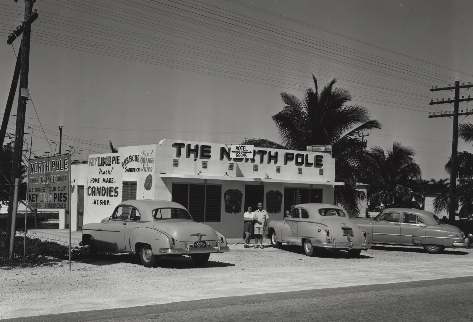 Black-and-white 1952 photograph of a flat-roofed roadside stand with 'The North Pole' in big letters and a 'Motel and Rooms' sign. Its walls are painted with 'Key Lime Pie Fresh!', 'Barbecue Sandwich', 'Fresh Orange Juice' and 'Home Made Candies - We Ship', and a freestanding sign at left reads 'North Pole - Fresh Orange Juice - We Make Our Own Key Lime Pies'. A man and a woman stand in the doorway, three early-1950s sedans are parked on the sand lot, and palms and utility poles rise behind under a clear sky.