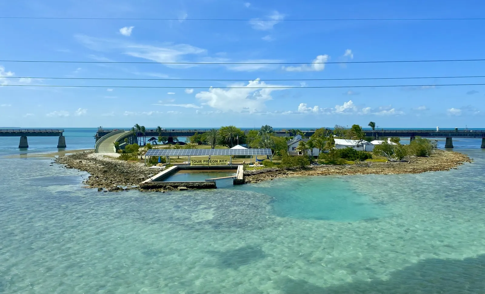 High view from the Seven Mile Bridge of Pigeon Key: a small low island ringed by rock and very clear turquoise shallows. A curved ramp rises from the island to the old steel-truss bridge that runs across the background on concrete piers. On the island are a solar-panel canopy over a sign reading 'Tour Pigeon Key', a small walled boat basin, white historic buildings, palms and lawns. Power lines cross the blue sky with scattered white cloud.