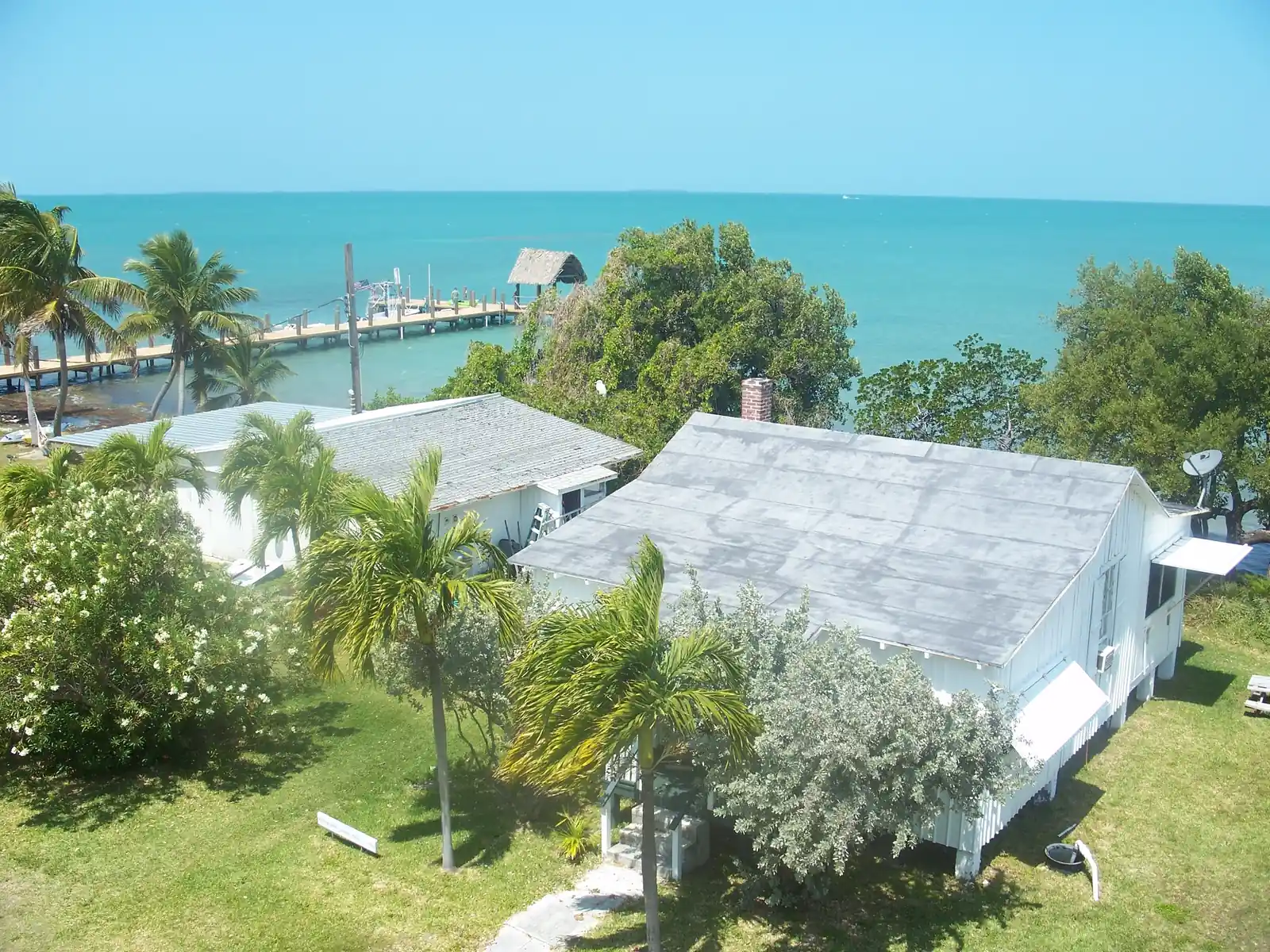 Elevated view over two white wooden historic buildings with grey roofs on Pigeon Key, one with a brick chimney and propped-open awning shutters, set on mown grass among palms, a white-flowering shrub and silvery buttonwood. Beyond them a long wooden pier with a thatched shelter and a few boats runs out into brilliant turquoise water that stretches to a flat horizon under a pale-blue sky.