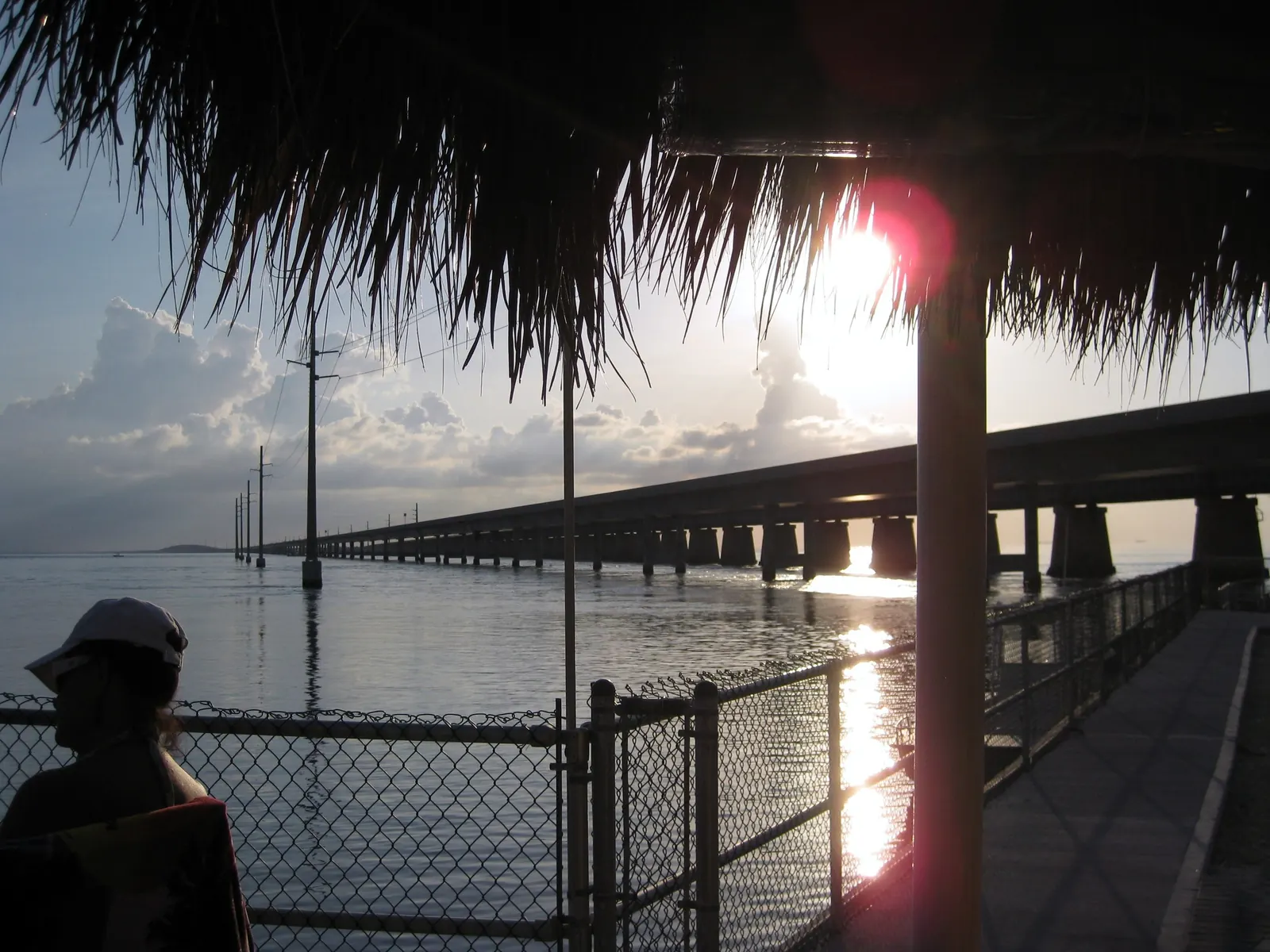 Strongly backlit view from under a thatched palapa on a fenced waterside deck: a long concrete highway bridge on tall piers runs from the right into the distance across calm, silvery water, with a line of utility poles standing in the water beside it. The low sun flares in red and white at upper right behind the thatch, with cumulus cloud on the horizon. A person in a cap is silhouetted at lower left.