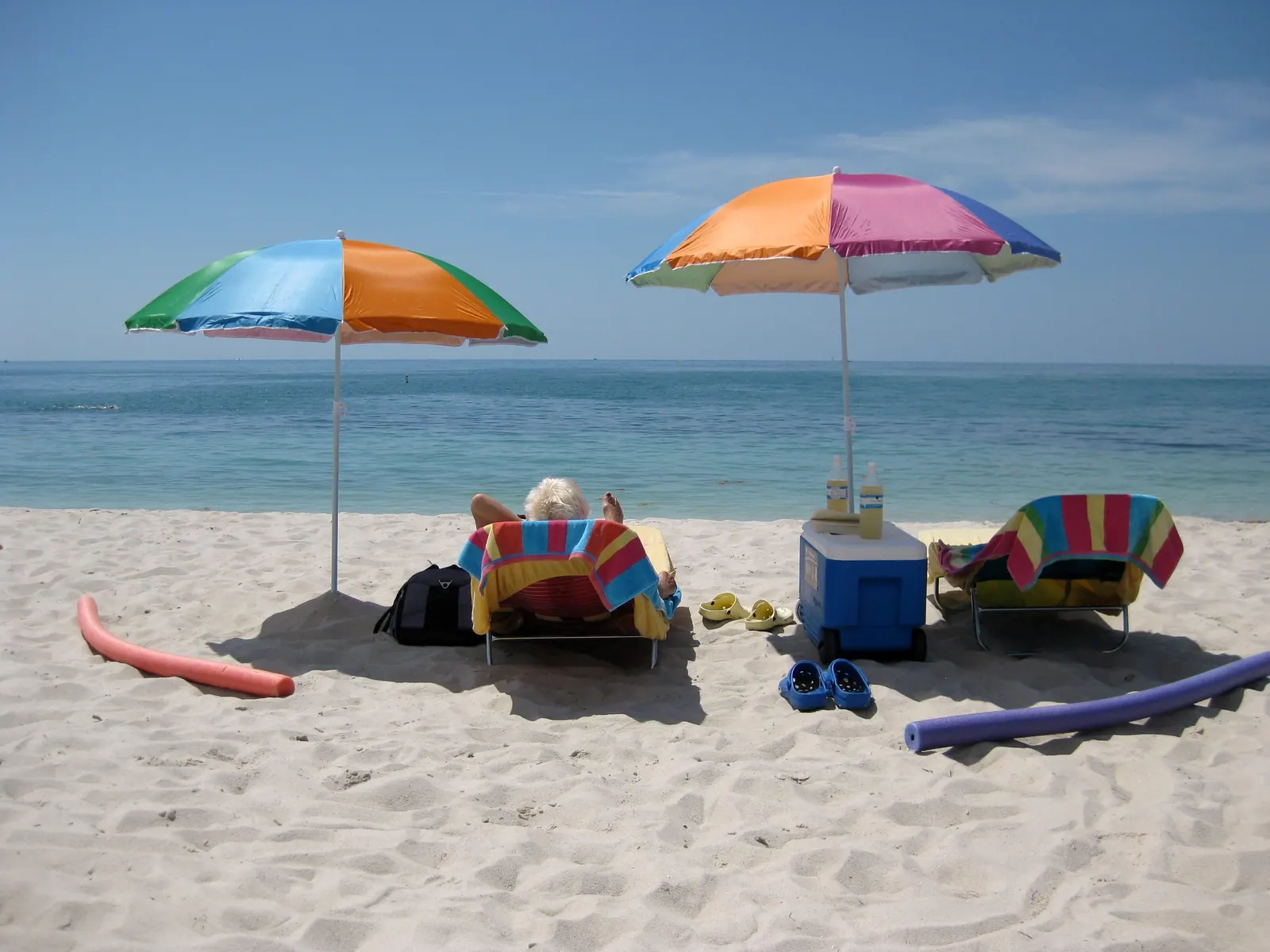 Two bright multicoloured beach umbrellas stand on pale, footprinted sand in front of calm turquoise water and a flat horizon under a pale-blue sky. Under the left umbrella a white-haired person lies back in a lounger draped with a striped towel, seen from behind; a second empty towel-draped lounger, a blue cooler with two drink bottles on top, pairs of plastic clogs, a black bag and pink and purple pool noodles lie around them.