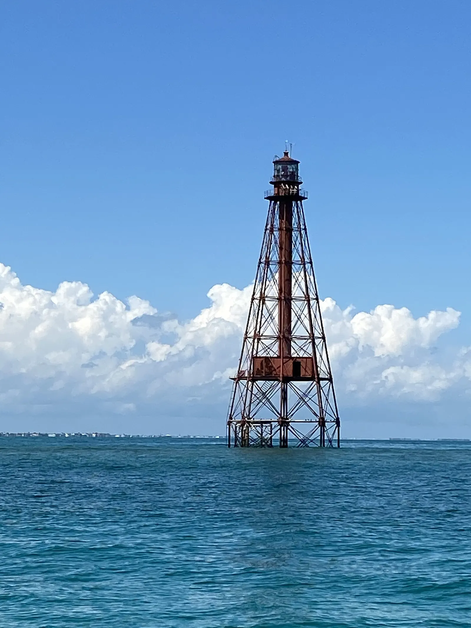 Vertical view of the rust-red iron skeleton tower of Sombrero Key Lighthouse rising from open blue-green water: tall tapering legs with cross-bracing, a central column, a boxy platform part-way up and a lantern room with a gallery at the top. Behind it is a bank of white cumulus and a deep-blue sky, with a thin line of low, distant shoreline on the horizon at left.
