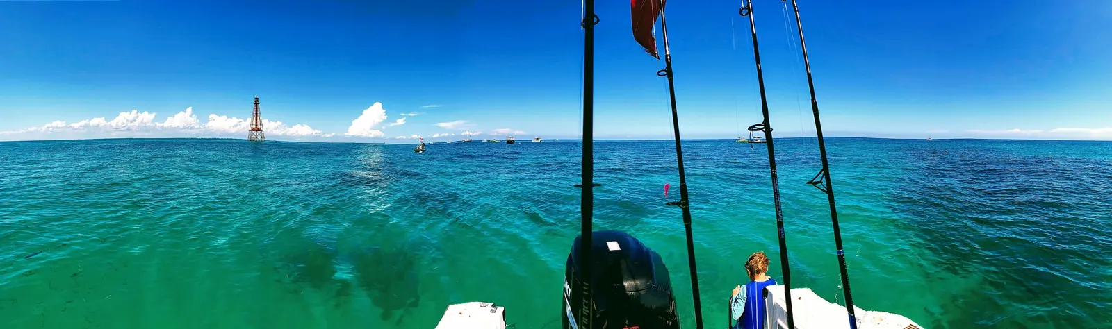 Wide phone panorama from the stern of a fishing boat over brilliant green-turquoise water above a reef, with dark patches of reef visible below the surface. The rust-red iron skeleton of Sombrero Key Lighthouse stands on the horizon at left in front of a bank of white cumulus, and small boats are moored across the middle distance. In the foreground are an outboard engine, four upright fishing rods and a child in a blue life vest seen from behind.