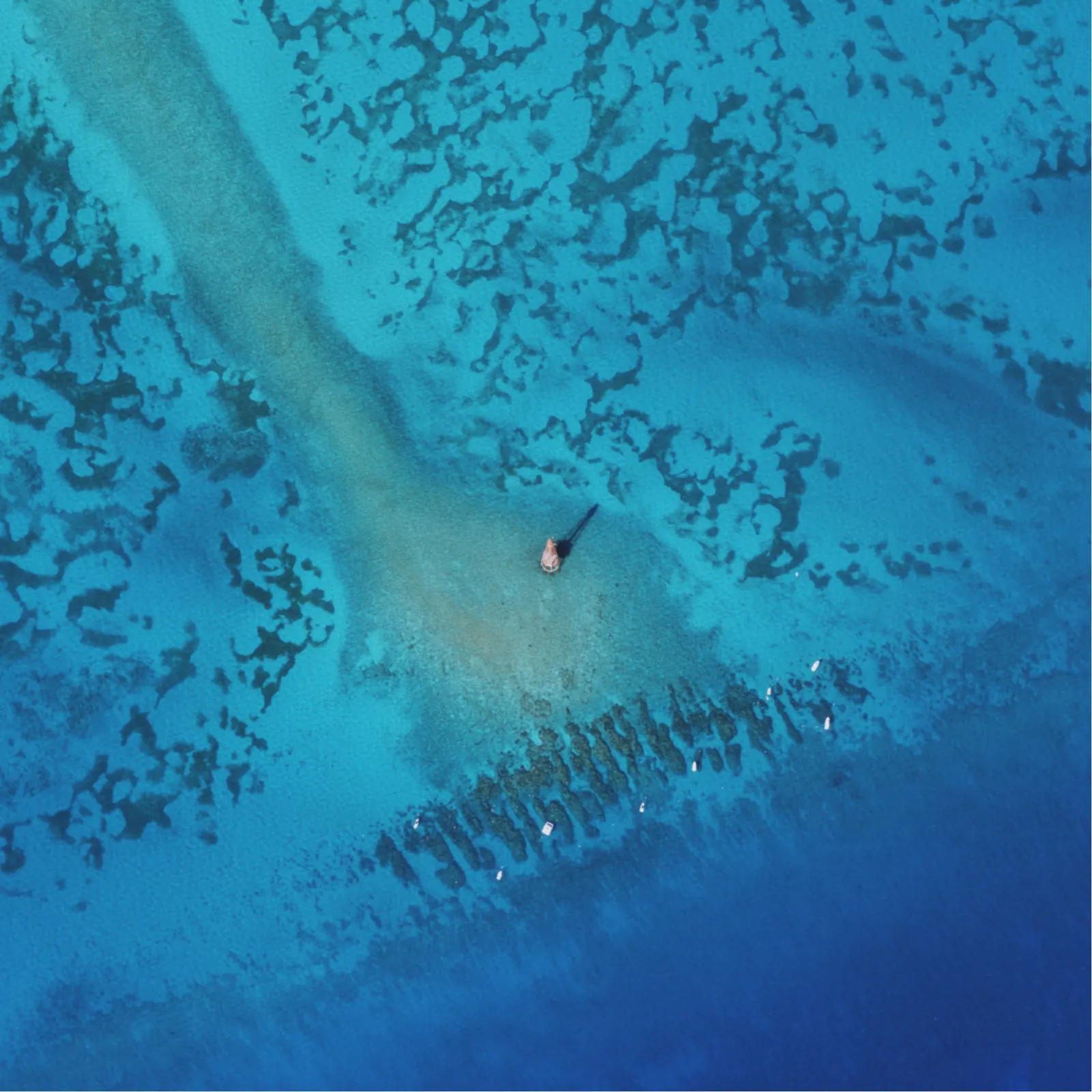 Straight-down aerial of the shallow reef: brilliant turquoise water with a pale sand tongue running diagonally to a small rust-coloured lighthouse structure at the centre, which casts a long shadow. Dark coral heads and patches cover the upper half, and at the bottom a row of dark finger-like spur-and-groove reef ridges runs into deep blue water, with about ten small white boats moored along it.