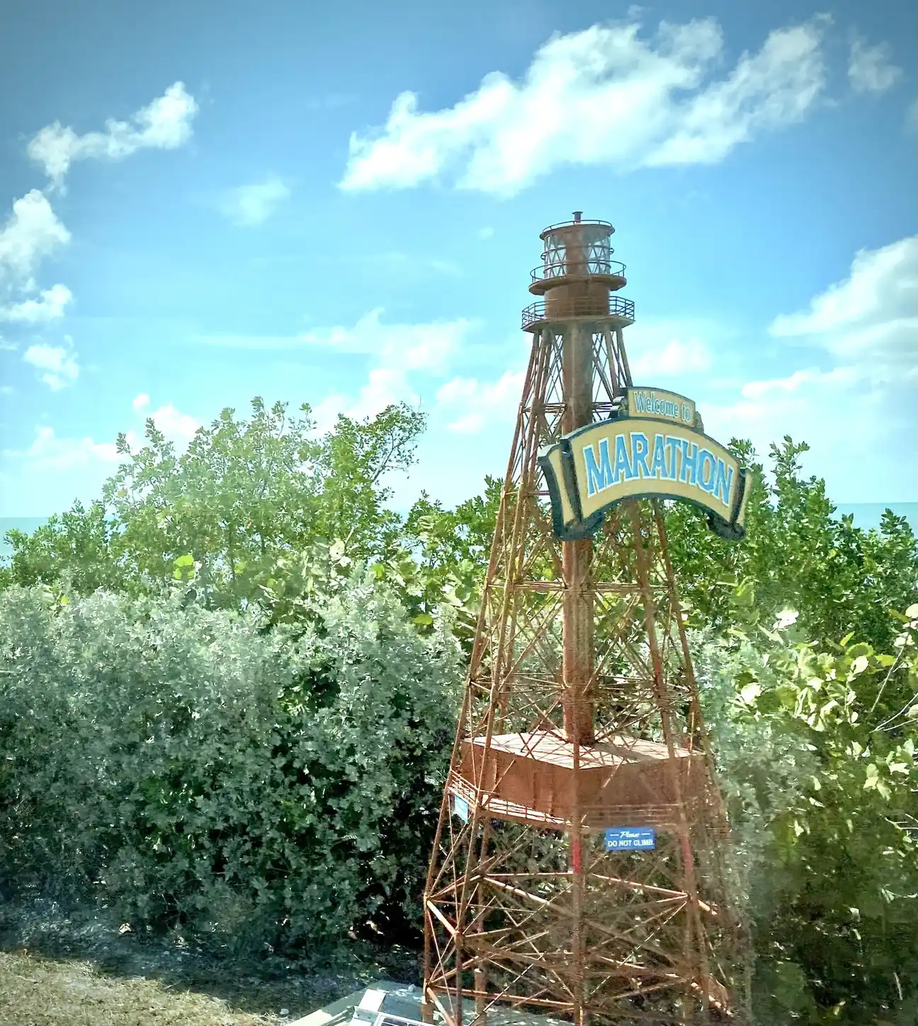 A rust-red iron skeleton-tower replica of a lighthouse, with a lantern room and railed galleries on top, carries an arched yellow-and-blue sign reading 'Welcome to Marathon'. It stands among dense green sea grape and silvery buttonwood shrubs with a glimpse of turquoise water behind, under a blue sky with wispy white cloud. A small blue plaque on the tower reads 'Please do not climb'.